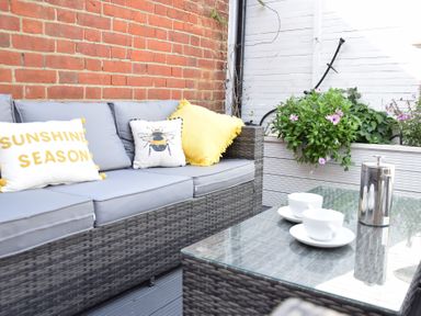 An outdoor seating area with a gray wicker sofa with cushions and decorative pillows and a glass coffee table with cups and a metal container at Regatta Lookout in Aldeburgh
