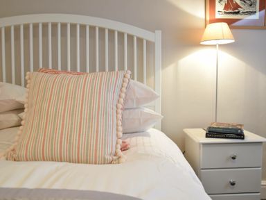 A bed with pillows and a striped cushion next to a white bedside table with books and a lamp at Regatta Lookout in Aldeburgh