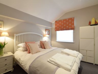 A bedroom with a white bed and pillows bedside tables lamps a window with orange patterned blind and a white cabinet at Regatta Lookout in Aldeburgh