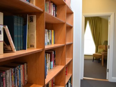 A wooden bookshelf filled with books and DVDs in a hallway leading to a room with an armchair and a lamp at Regatta Lookout in Aldeburgh