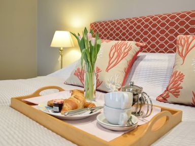 A wooden tray with croissants jam coffee cups and flowers on a bed with patterned cushions and a lamp at Regatta Lookout in Aldeburgh
