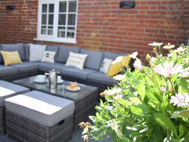 An outdoor patio with a gray sectional sofa with cushions a glass coffee table with a coffee pot and cups and flowers in the foreground at Regatta Lookout in Aldeburgh