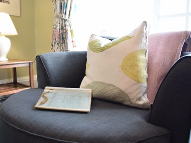 A dark gray armchair with patterned and pink pillows a book on the seat next to a wooden side table with a white lamp at Regatta Lookout in Aldeburgh