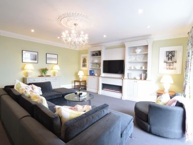 A living room with a large dark sectional sofa a round glass coffee table a chandelier and a wall-mounted television at Regatta Lookout in Aldeburgh