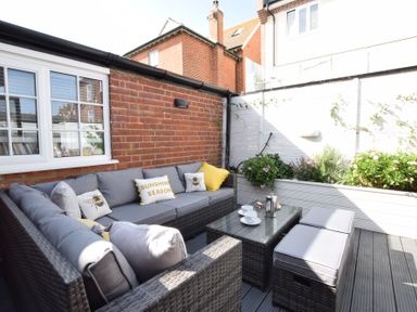 An outdoor seating area with a grey wicker sofa set and cushions beside a brick wall with plants in white planters at Regatta Lookout in Aldeburgh