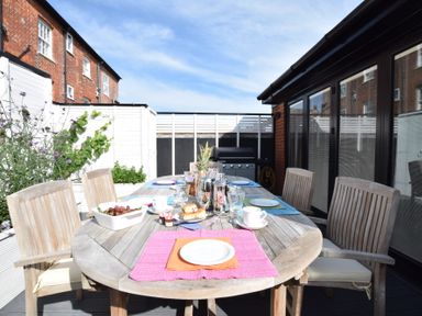 An outdoor dining table with chairs plates cups and a bowl of grapes on a terrace with a barbecue grill at Regatta Lookout in Aldeburgh