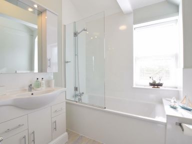 A bathroom with a white sink and cabinet a bathtub with a glass shower screen and a window with model boats on the sill at Regatta Lookout in Aldeburgh