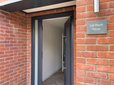 An open black door with a metal handle set in a brick wall next to a sign reading Salt Marsh House in Southwold