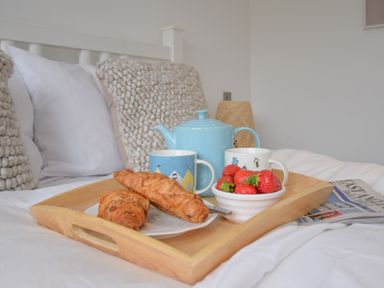 A wooden tray with a teapot cups croissants strawberries and a newspaper on a bed with pillows at Salt Marsh House Southwold