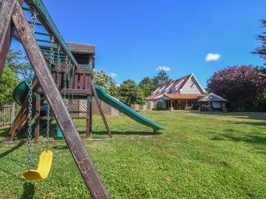 A garden with a children's playset that has a yellow swing and a green slide next to a house with a red tiled roof and a covered terrace at Moat Barn Hasketon Near Woodbridge