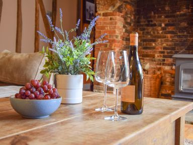 A wooden table with a bowl of grapes a plant in a pot a wine bottle and two wine glasses in front of a brick fireplace at Moat Barn in Hasketon Near Woodbridge