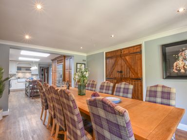 A dining area with a wooden table and plaid upholstered chairs next to a kitchen with white cabinetry and wooden floor at Moat Barn Hasketon Near Woodbridge