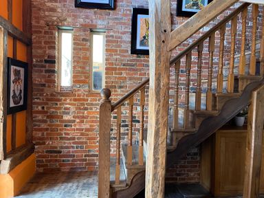 A wooden staircase with spindles next to a brick wall with small vertical windows and framed pictures at Moat Barn in Hasketon Near Woodbridge
