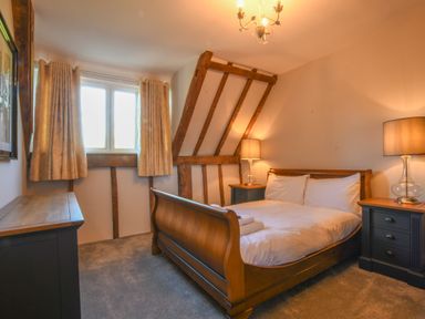 A bedroom with a wooden bed flanked by two bedside tables with lamps and a window with curtains at Moat Barn in Hasketon Near Woodbridge