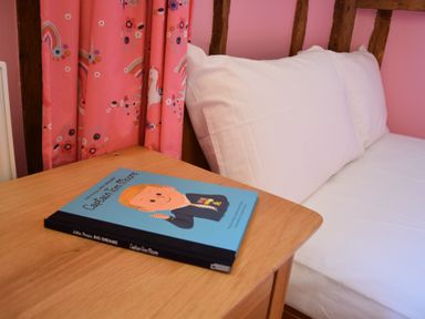 A wooden bedside table with a children's book next to a bed with white pillows and pink curtains with unicorns at Moat Barn Hasketon Near Woodbridge