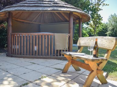 A wooden bench with a bottle and glass on a patio near a thatched gazebo in a garden at Moat Barn Hasketon Near Woodbridge