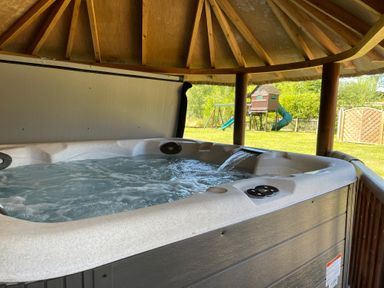 A hot tub with water flowing inside a wooden gazebo with a children's playset in the background at Moat Barn in Hasketon Near Woodbridge
