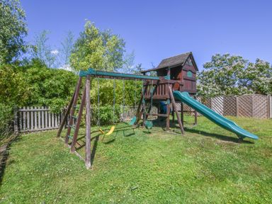 A wooden outdoor playset with swings slide and climbing area in a fenced grassy garden at Moat Barn in Hasketon Near Woodbridge
