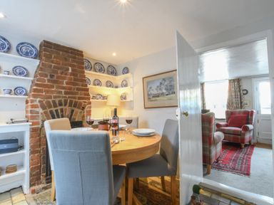A dining room with a wooden round table set with plates and wine glasses a brick fireplace and shelves with blue and white plates at 17 South Green Southwold