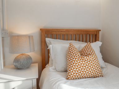 A bed with white sheets and a patterned pillow next to a nightstand with a lamp at 17 South Green Southwold