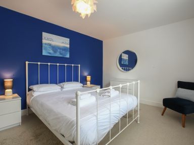 A bedroom with a white metal bed frame a blue accent wall two bedside tables with lamps a round wall mirror and a dark chair at The Gallery Blythburgh near Southwold