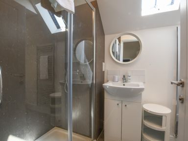 A bathroom with a glass shower enclosure a white sink with cabinet under a round mirror and a small white shelving unit at The Gallery Blythburgh near Southwold