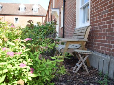 A wooden bench and small wooden stool next to a brick wall with green plants and pink flowers in the foreground at The Gallery  Blythburgh