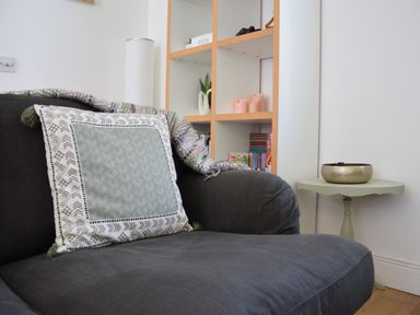 A dark grey armchair with a patterned cushion and a throw blanket next to a wooden shelving unit and a small round side table with a brass bowl at The Gallery Blythburgh near Southwold