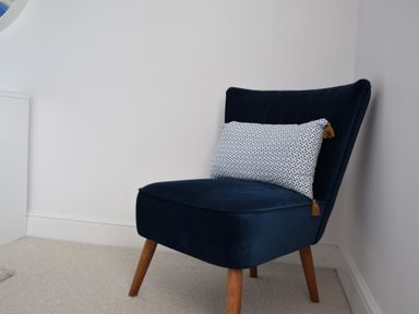 A blue upholstered chair with wooden legs and a patterned rectangular cushion in a corner of a white room at The Gallery Blythburgh near Southwold