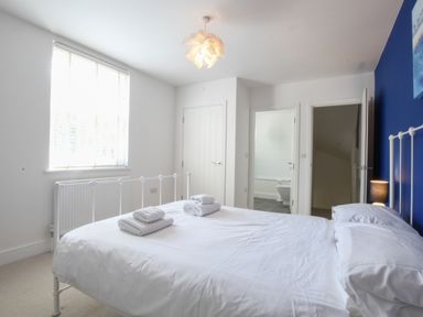 A bedroom with a white metal bed frame white bedding folded towels on bed a blue accent wall and doors to bathroom and hallway at The Gallery Blythburgh near Southwold