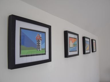 Four framed pictures with colorful building illustrations on a white wall at The Gallery Blythburgh near Southwold