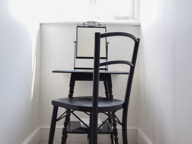 A small black table with an iron-framed mirror and a black chair in a narrow white room at The Gallery Blythburgh near Southwold