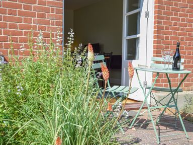 A garden area with green plants and flowers next to a brick wall with a small green table and two chairs with wine glasses and a wine bottle at The Gallery Blythburgh near Southwold