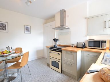 A kitchen with wooden countertops, cream cabinets, a stove, a microwave, a toaster, and a round glass dining table with wooden chairs with fruit and croissants at Driftwood Southwold