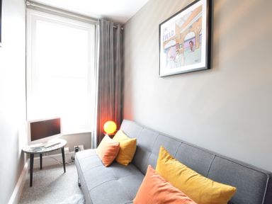A small living room with a grey sofa with orange and yellow cushions a side table with a television and an orange lamp and a framed picture on the wall at Driftwood Southwold in Southwold