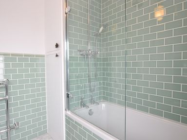 A bathroom with a bathtub and a shower next to a white storage cabinet with green tiled walls at Driftwood Southwold