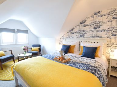 A bedroom with a bed with yellow and blue bedding two chairs with yellow cushions and a small round table with flowers at Driftwood Southwold