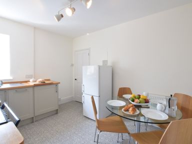 A kitchen with wooden chairs around a glass table with fruit and pastries a white refrigerator and cabinets at Driftwood Southwold