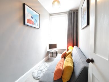A narrow room with a gray sofa with orange and yellow pillows a small table with a monitor a furry rug and framed pictures on the walls at Driftwood Southwold