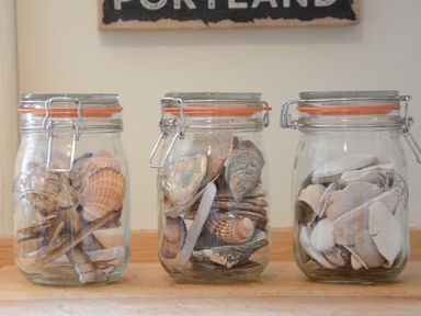 Three glass jars filled with various seashells placed on a wooden surface beneath a wall hanging