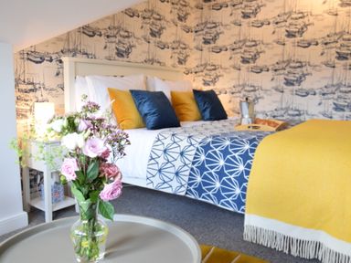 A bedroom with a bed with blue and yellow pillows and a yellow blanket a bedside table with a lamp and a vase with flowers on a round table at Driftwood Southwold