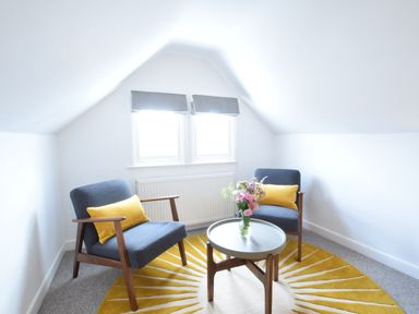 A small sitting area with two blue chairs with yellow cushions and a round table with flowers on a yellow and white rug at Driftwood Southwold