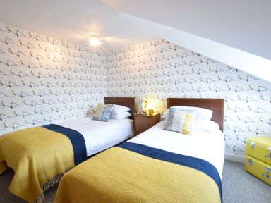 A bedroom with two single beds yellow throws and bird patterned wallpaper at Driftwood Southwold