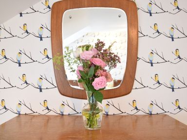 A vase with pink flowers on a wooden table in front of a wall mirror with bird wallpaper at Driftwood Southwold