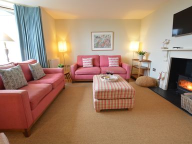 A living room with pink sofas a plaid ottoman a fireplace and blue curtains at Tamarisk in Aldeburgh