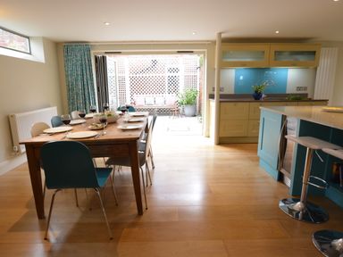 A dining area with a wooden table set for six people next to an open kitchen and patio at Tamarisk Aldeburgh in Aldeburgh