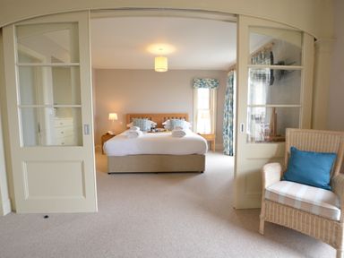 A bedroom with a large bed and side tables seen through sliding glass doors with a wicker chair in the foreground at Tamarisk Aldeburgh in Aldeburgh