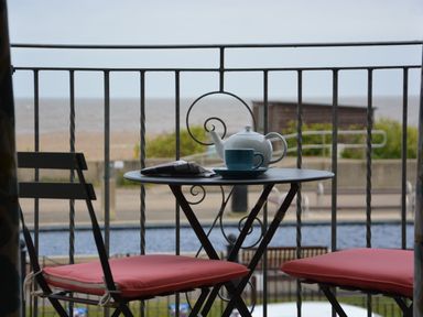 A small balcony with a black metal table and two chairs with red cushions overlooking a beach with a teapot cup and newspaper on the table at Tamarisk Aldeburgh in Aldeburgh