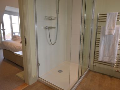 A bathroom with a glass shower enclosure white towels on a heated towel rail and a bedroom visible through an open door at Tamarisk Aldeburgh