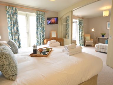 A bedroom with a bed with white linens and rolled towels a wooden tray with breakfast a TV on the wall floral curtains and an adjacent sitting area with chairs and a table at Tamarisk Aldeburgh